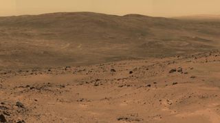 Martian landscape view from NASA's Mars rover Spirit, showing a vast, rocky terrain with gentle hills in the distance. The scene has a reddish-brown hue typical of the Martian surface, with scattered rocks and a dusty, barren ground stretching to the horizon.