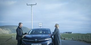 Sandy (Steven Robertson) and Calder (Ashley Jensen) stand on either side of a car parked in a passing place, both leaning on the bonnet. Calder looks exasperated and emotional.