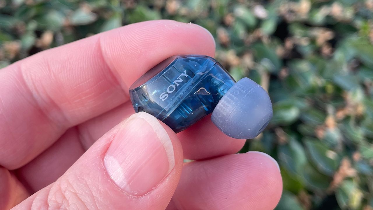A hand holding a transparent blue Sony WF-C710N in-ear headphone.