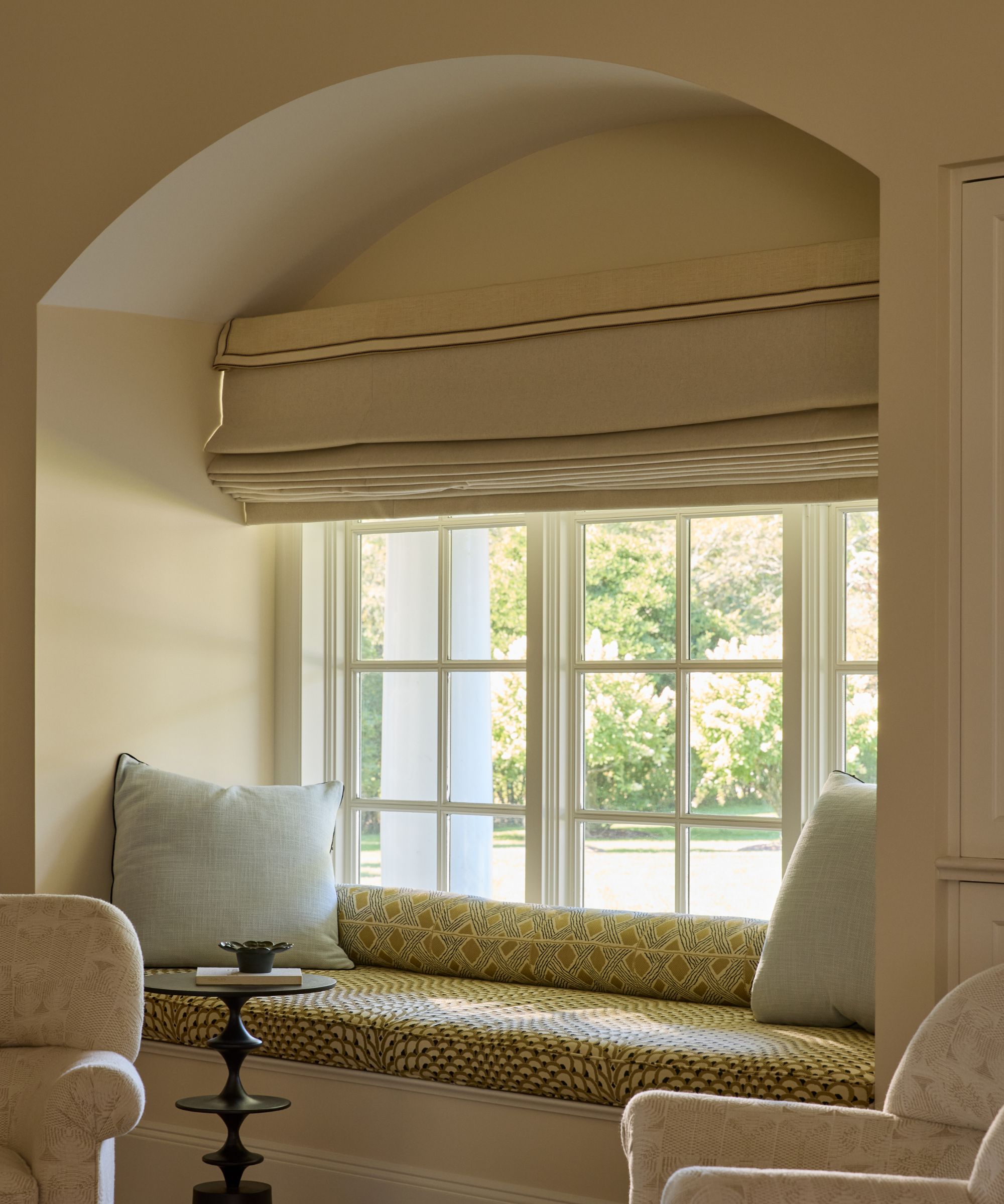 A built-in window seat beneath a tailored linen Roman shade with contrast piping, framed by an arched alcove and white-painted windows overlooking the garden.
