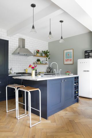 Corner of kitchen with blue Shaker-style island, wood and white metal bar stools, herringbone floor, glass pendants overhead and pale green rear wall