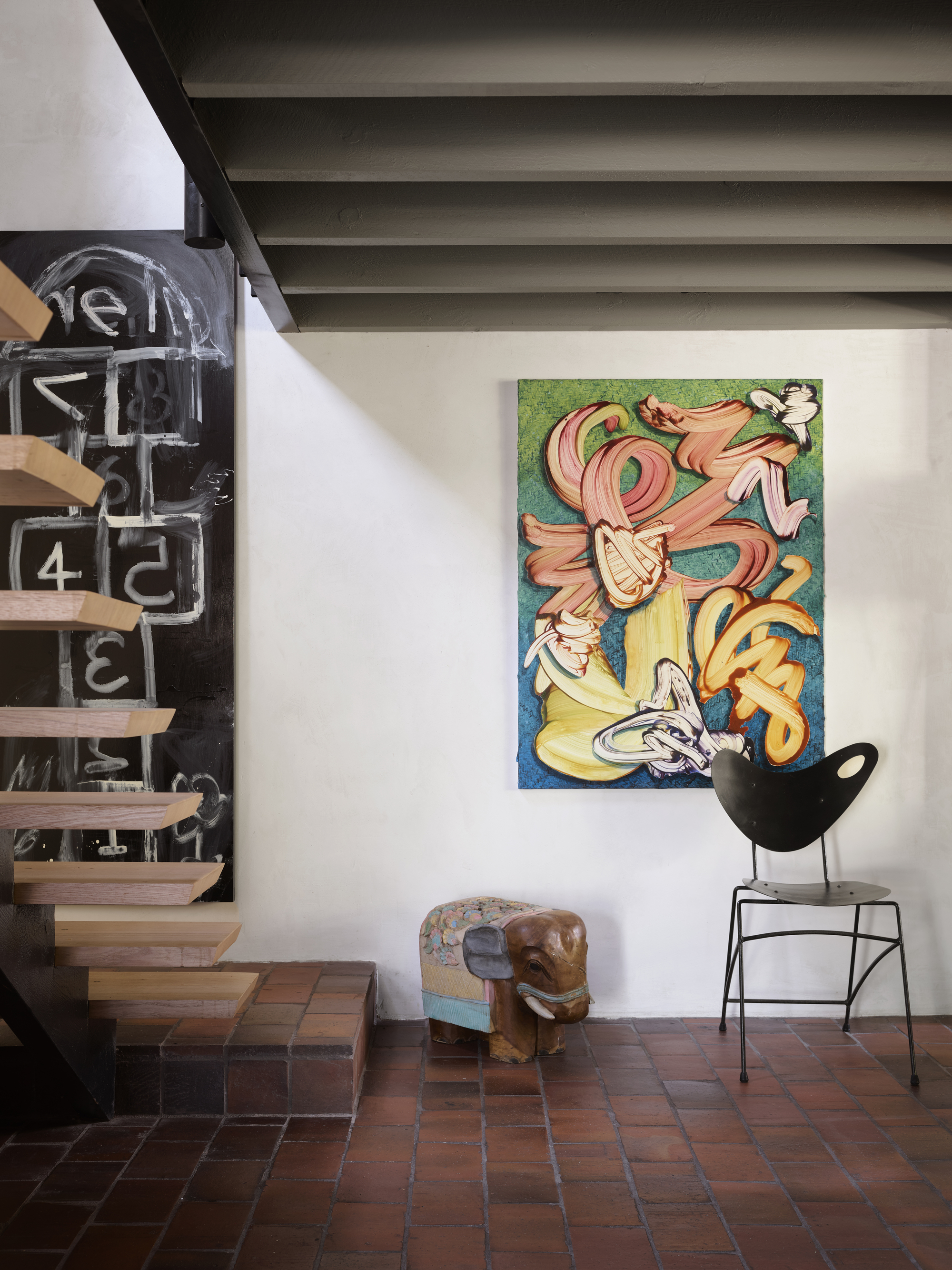 entryway of modern home with terracotta floor tiles, exposed beam ceiling painted green, timber floating staircase, artwork on walls, chair and sculpture of elephant
