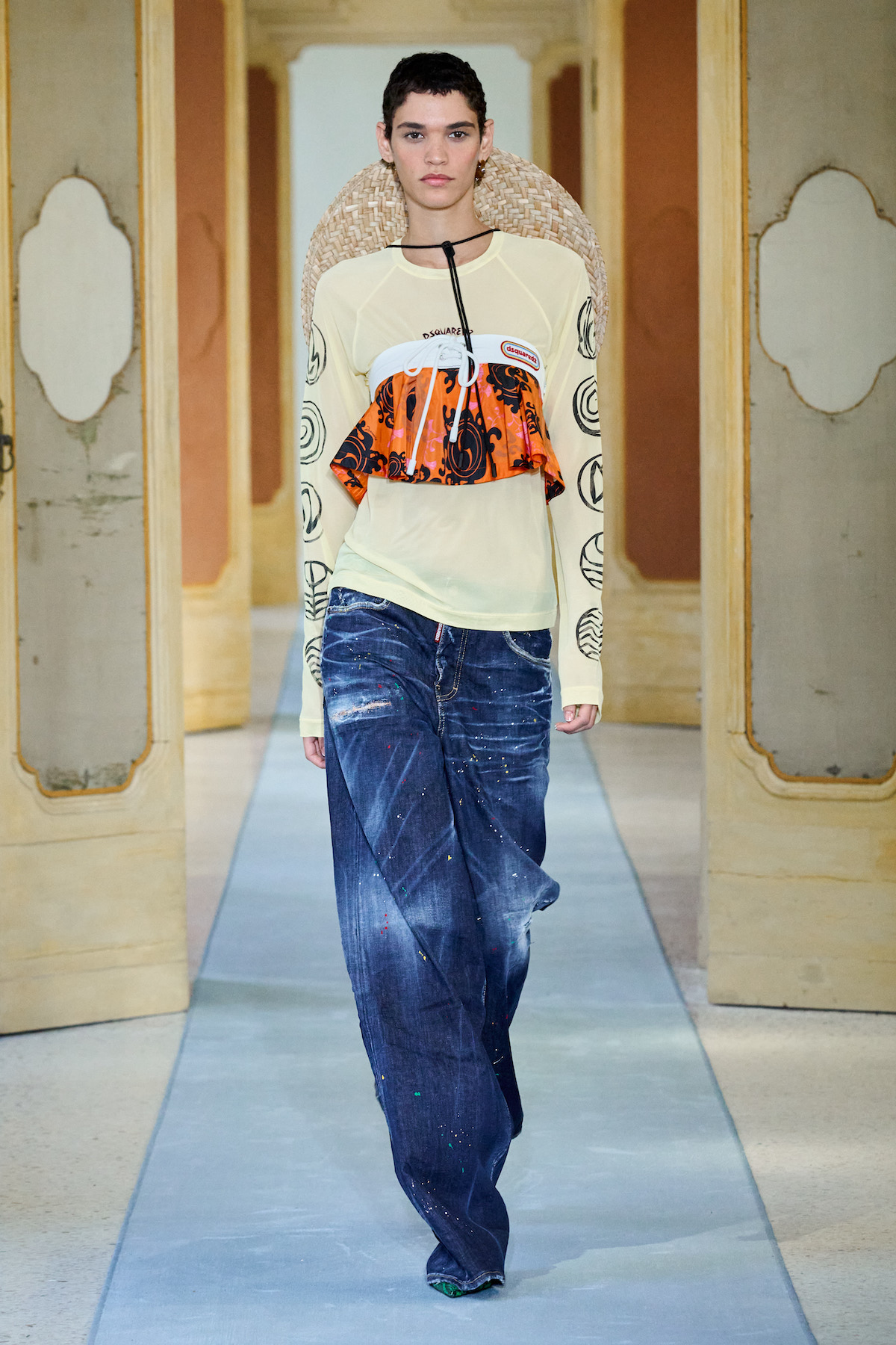 A female model wearing a long sleeve beige shirt, an orange bikini over it, baggy blue jeans and a large hat.
