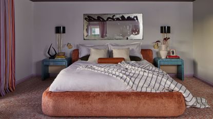 A dimly lit bedroom with a mirror above the bed, with a dusty rose textured holder and headboard with blue bedside tables and matching wall sconces