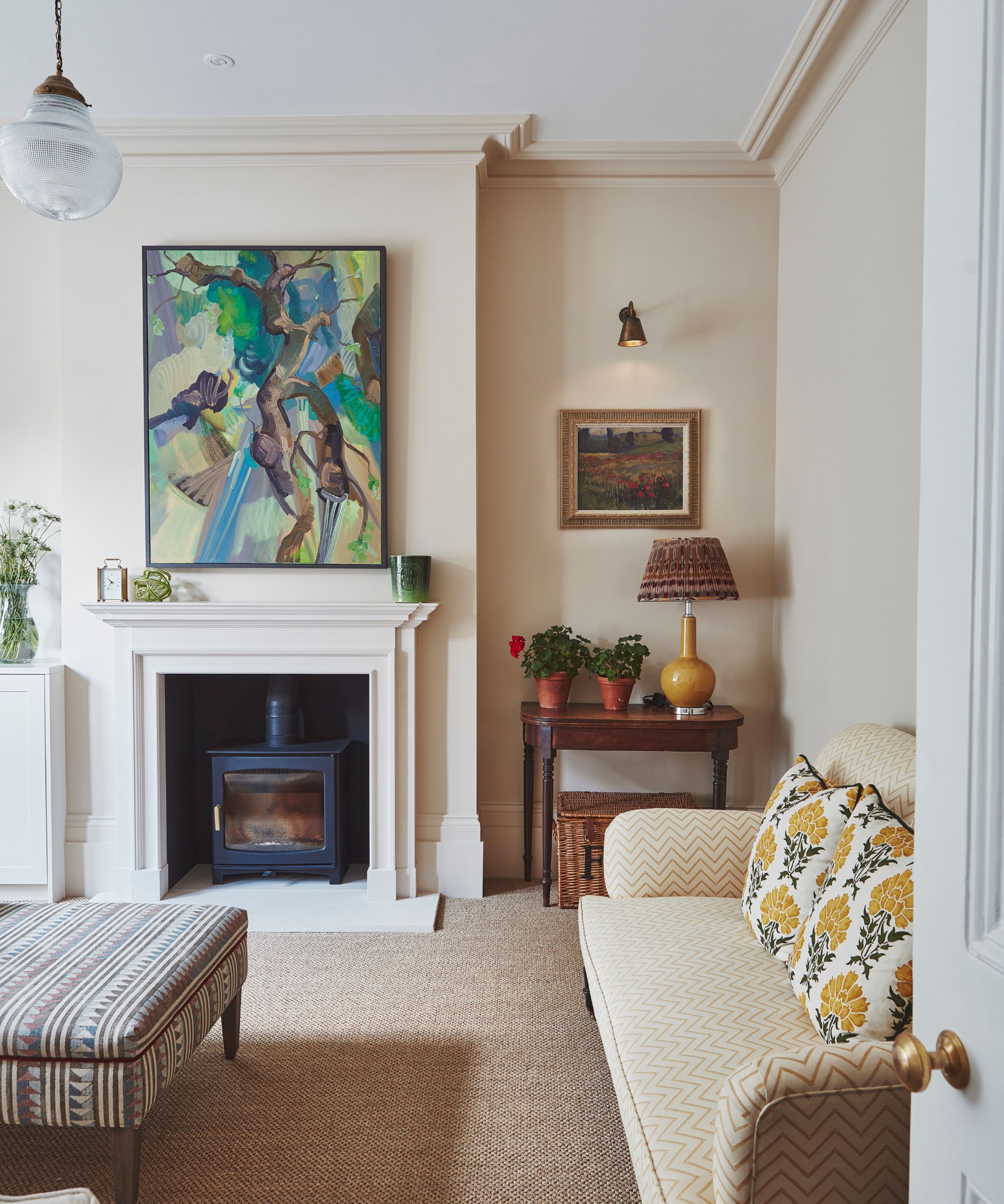 Traditional living room with cream painted walls, rattan carpet, yellow chevron sofa, ottoman, wood burner, abstract art above the fireplace and a nook with a framed painting, antique wooden console and yellow lamp underneath