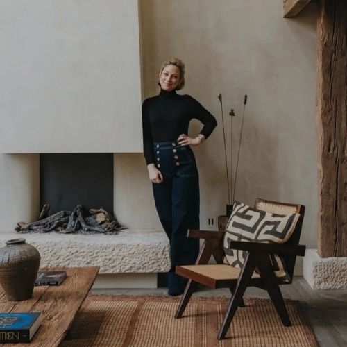 Image of a woman in a black turtleneck and dark jeans standing in a living room with a rustic, modern aesthetic. She's leaning against a stone fireplace.
