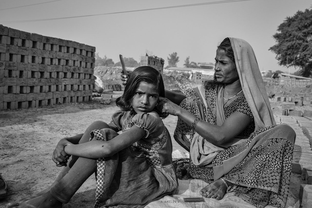 A black and white image showing girl and a woman sitting with each other, wearing tradidional clothing, the woman takes care of the girl's hair