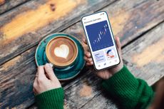 A person's hands holding a coffee with a heart latte art and their smartphone displaying a banking app with a graph increasing steadily.