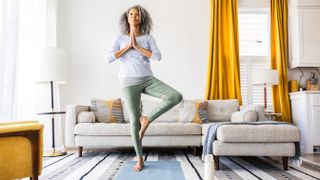 A woman stands practicing tree pose in her living room. She is standing on her right leg, with her left leg bent and left foot planted next to her right knee. Her hands are clasped together at her chest. Behind her is a couch, lamp, and large window framed by bright curtains.