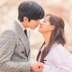 a man in a plaid suit and a woman in a purple top lean in for a kiss in a still from the k-drama business proposal