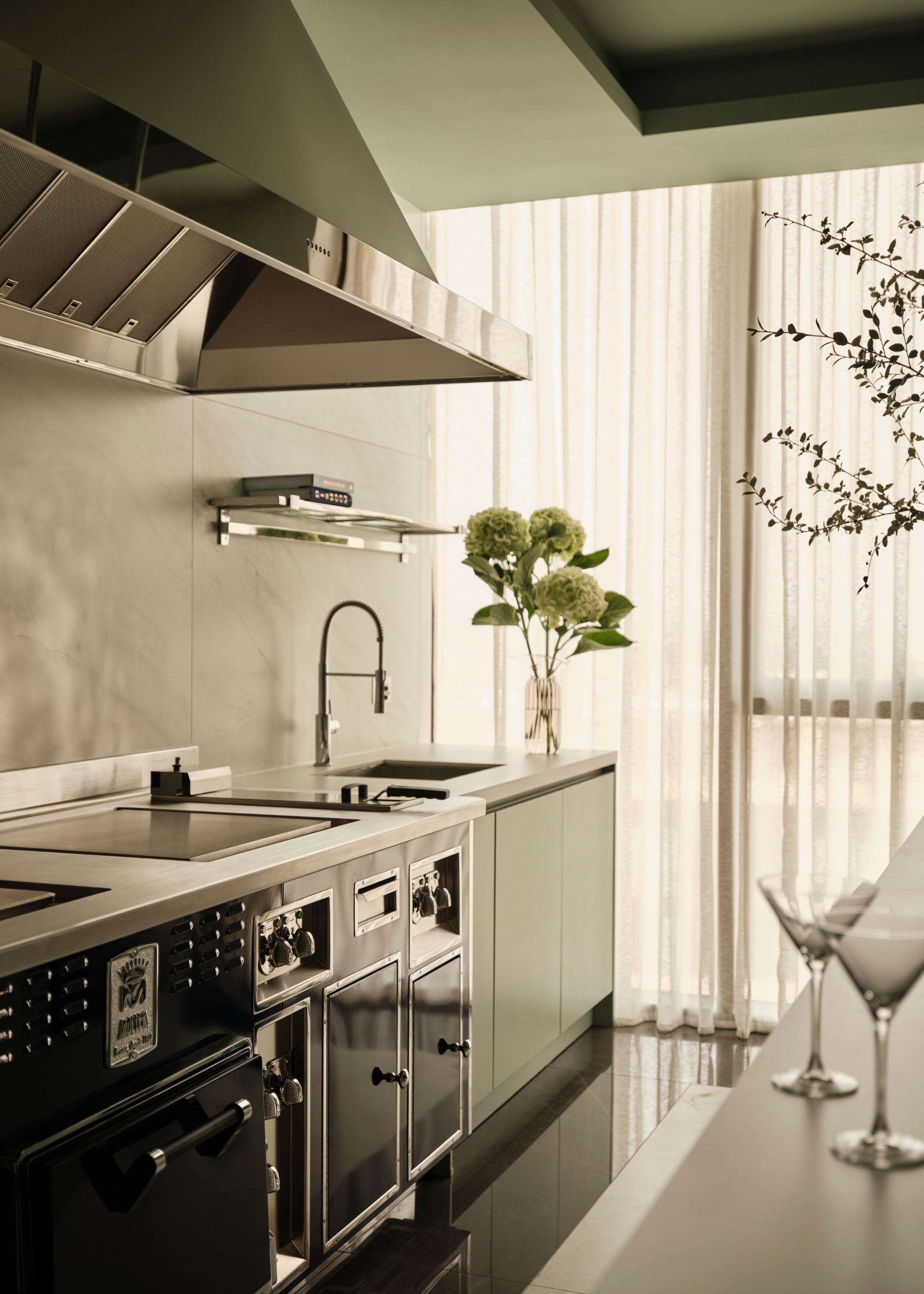 A modern galley kitchen with stainless steel and chrome accents with a hydrangea arrangement in a vase on the work surface and two emoty cocktail glasses with sheer curtains pulled over the window