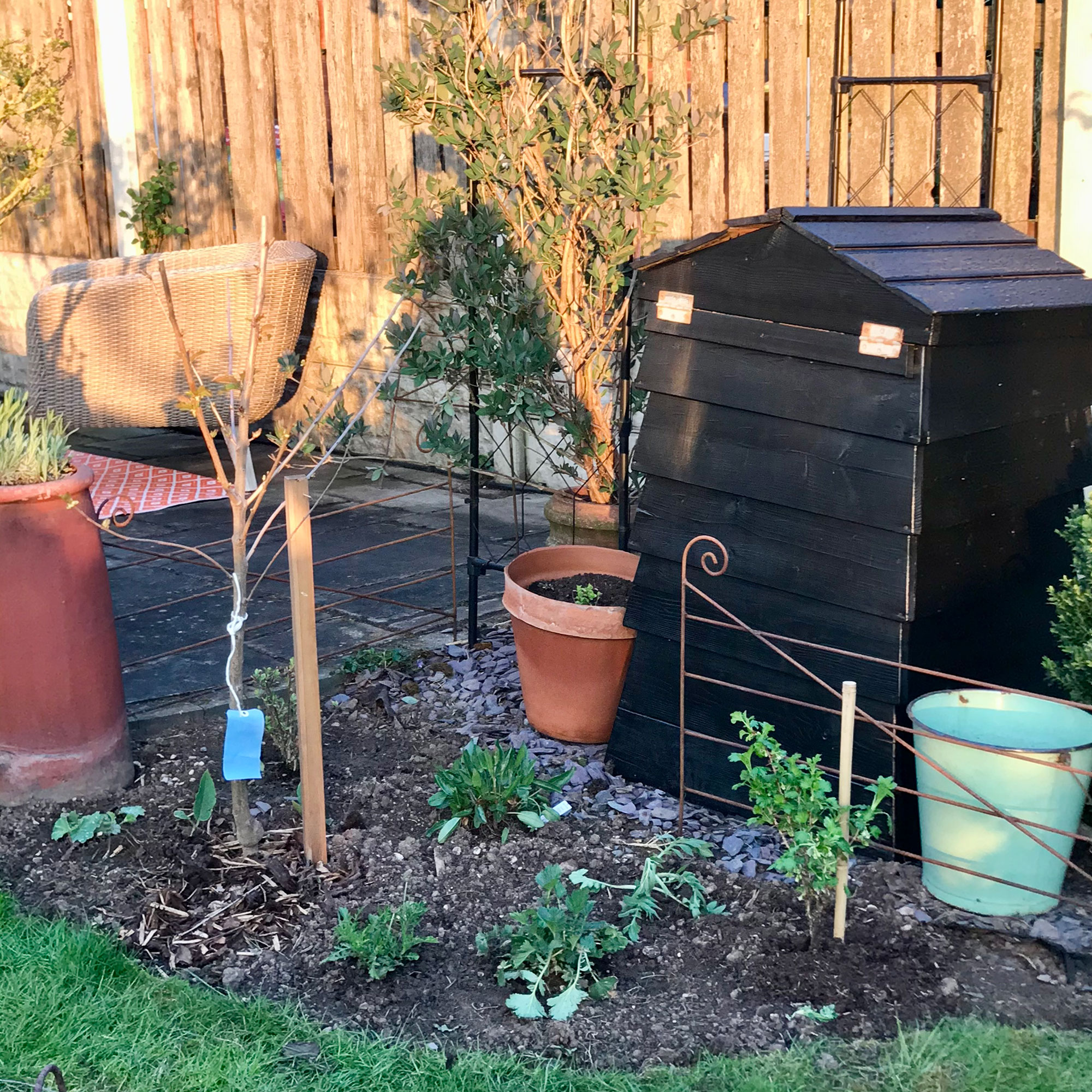 Black shed-like compost bin in garden by flower bed, wooden fence and patio area with seating