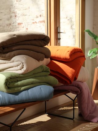 Brooklinen Airweave quilts in different colors placed on top of a bench. The quilt colors are orange, blue, khaki, green, cream and blush pink.