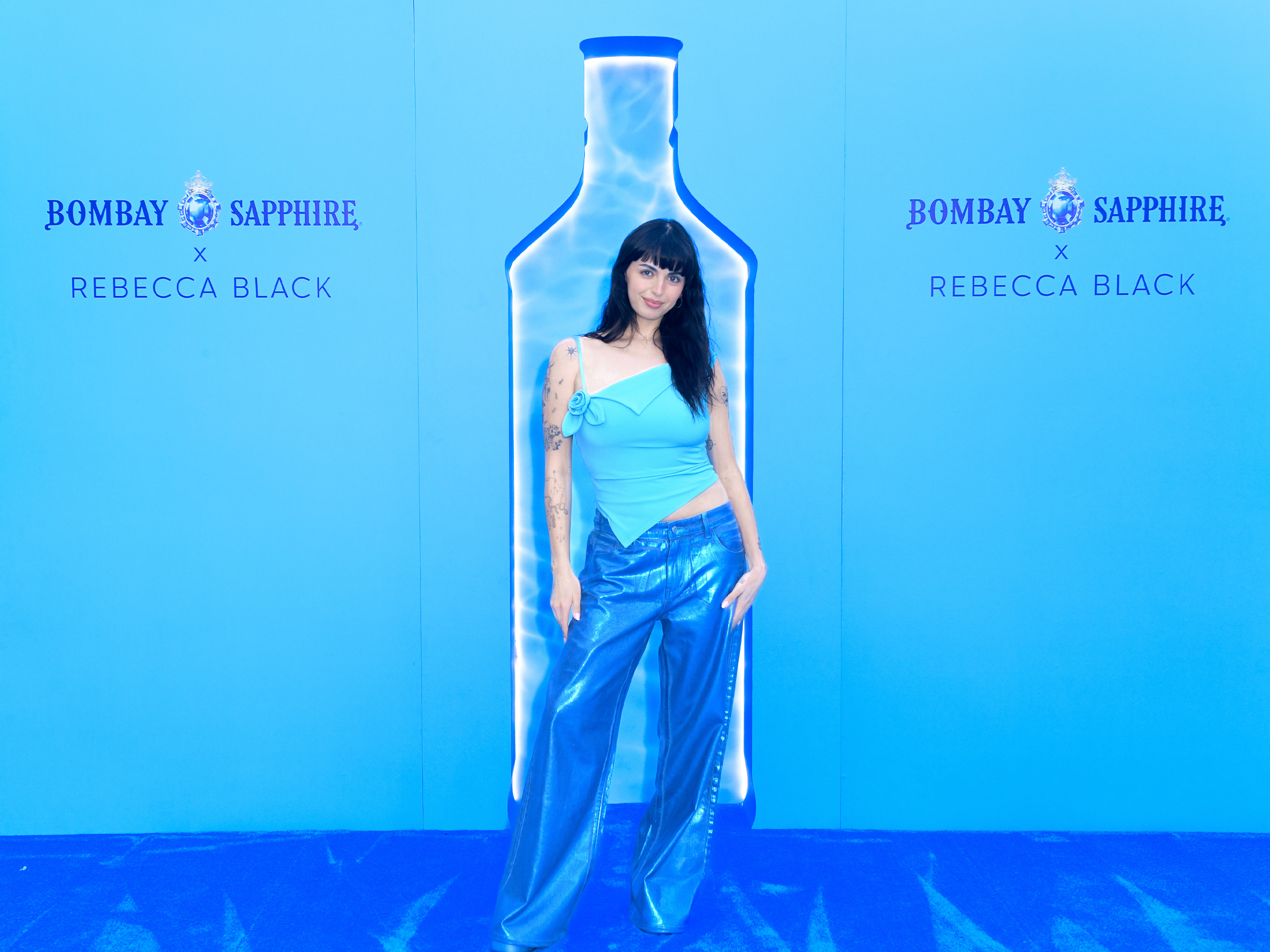 Singer Rebecca Black is standing in front of a large blue wall with a silhouette of a Bombay Sapphire gin bottle on it. The wall also has 