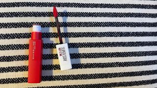 An open tube of Maybelline Superstay Matte Ink Lipstick in a bright red shade, on a striped background, for the purpose of the Maybelline Superstay Ink Review