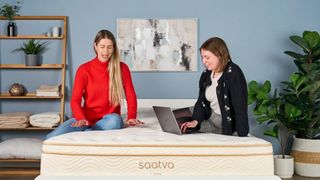 The image shows two mattress sat on top of the cream and gold Saatva Classic mattress. One tester is sitting on the bed while typing onto a laptop while the other places her hands on the surface of the bed.