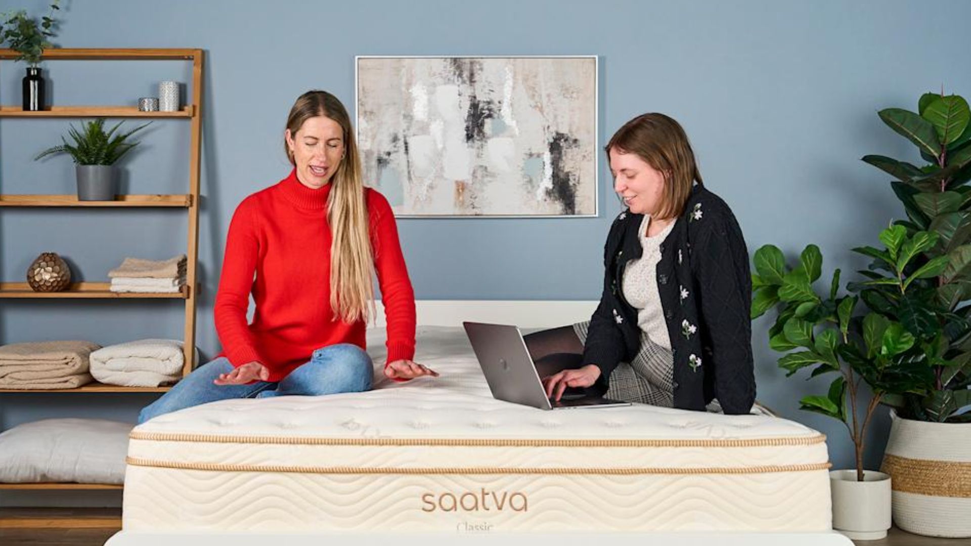 The image shows two mattress testers sat on top of the cream and gold Saatva Classic mattress. One tester is sitting on the bed while typing onto a laptop while the other places her hands on the surface of the bed.
