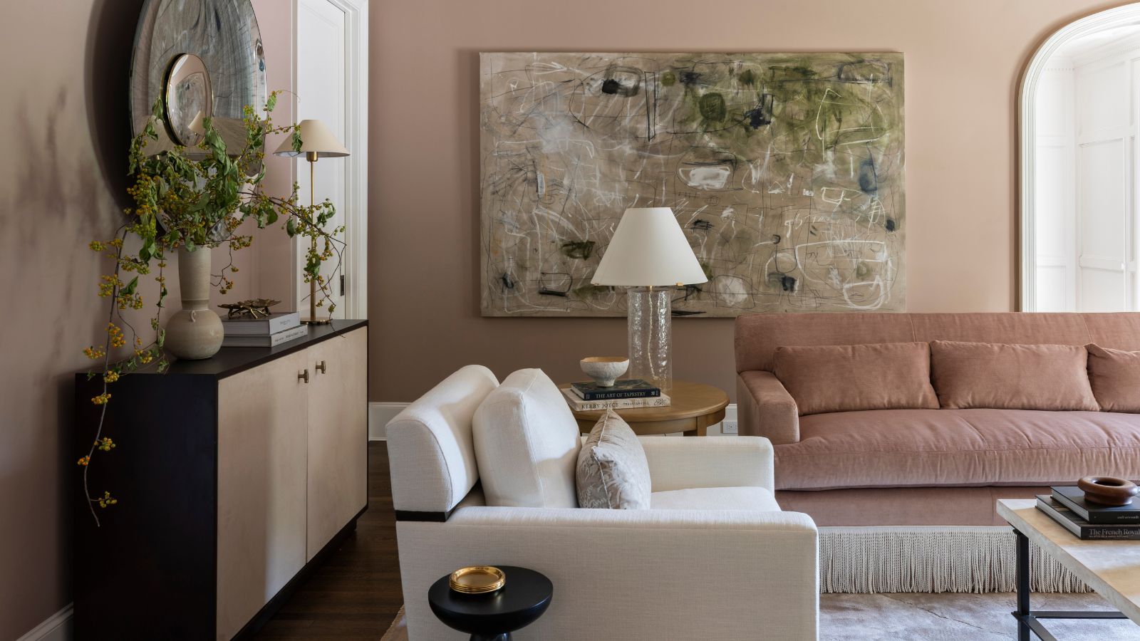 blush pink living room with a dusty pink velvet fringe sofa, white armchair, a large sideboard with artwork, and an oversized abstract painting on the back wall