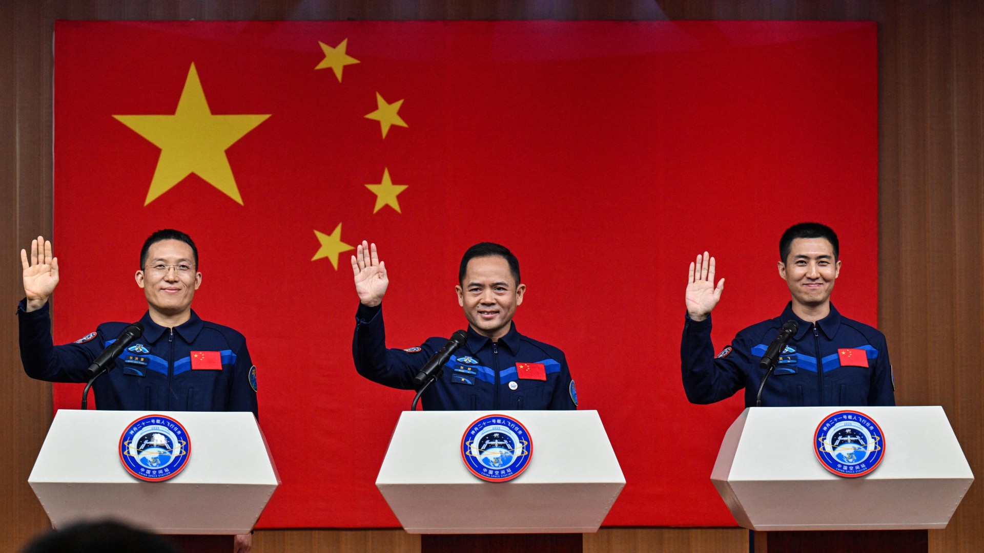 Three men wearing uniforms stand behind three podiums with a Chinese flag behind them