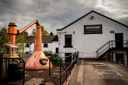 A copper whisky still in front of a white distillery building with a grey sky