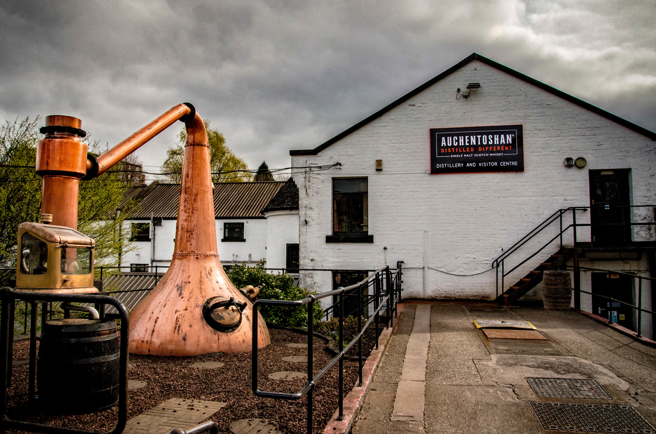 A copper whisky still in front of a white distillery building with a grey sky