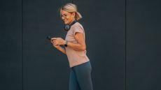 A woman walks in leggings and a t-shirt, looking at her phone and wearing over-ear headphones.