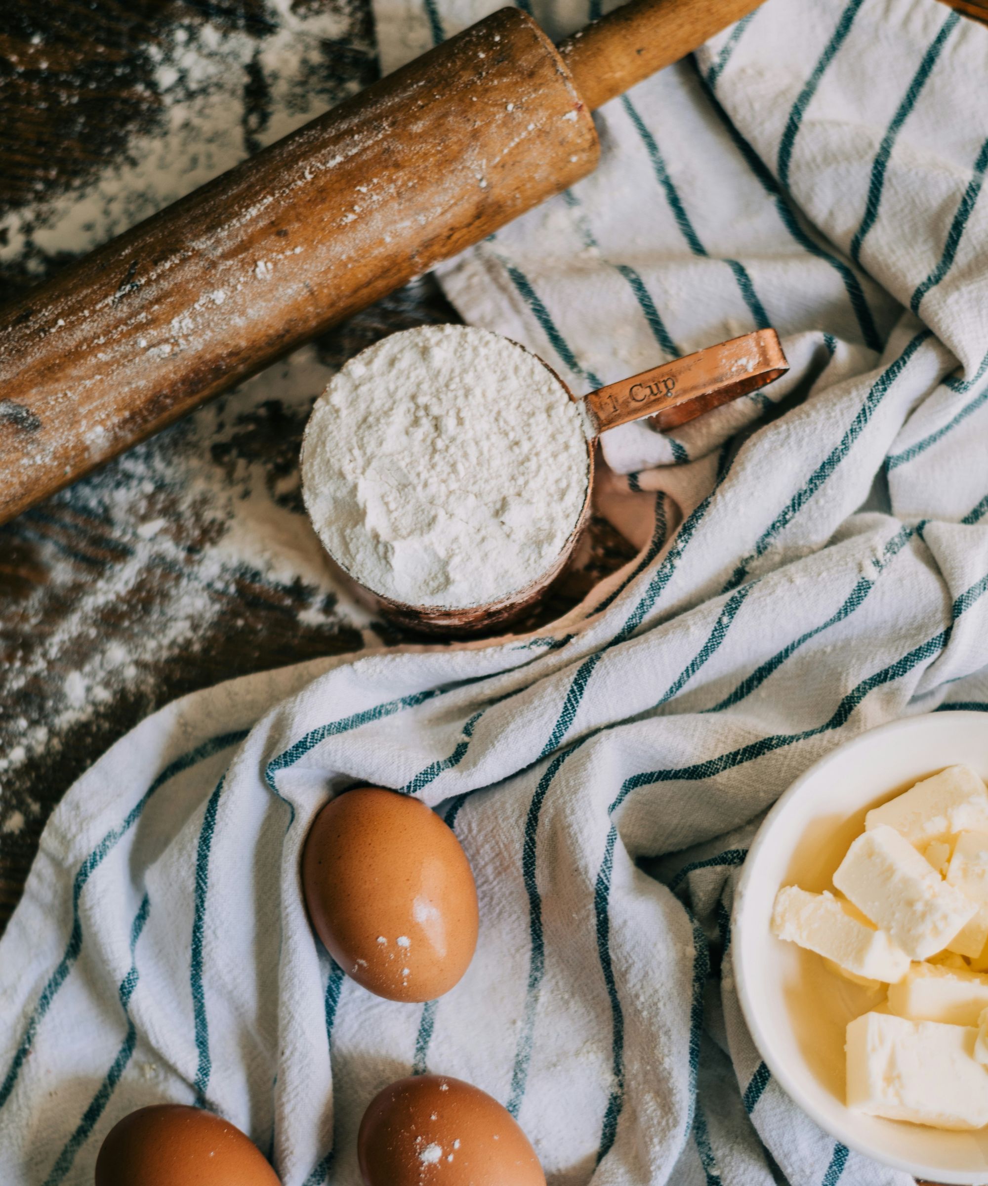 Eggs, butter and flour on top of blue and white striped tea towel next to wooden rolling pin