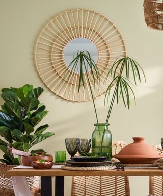 A pale green wall with a rattan circular mirror with a light brown wooden dining table in front of it with green and gray glassware, plates tall plants, a terracotta pan on it