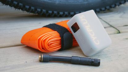 A tubolito TPU inner tube and Cycplus AS2 Ultra electric pump with its valve extension placed on a wooden deck, with a gravel tyre in the background