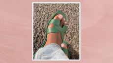 A close-up of someone's feet with a French tip pedicure, by nail artist, Julia Diogo (@paintedbyjools), and wearing green sandals, in front of a pavement background and in a pink template