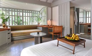 Surin ensuite hotel room with built in sofa with beige cushions, grey wood panelling, white linen with wooden lap try of fruit on bed