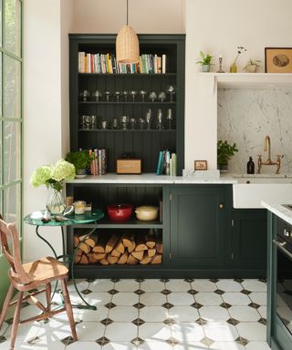 Dark green kitchen cabinets with white walls, geometric and retro looking diamond patterned floor tiles, and logs stacked in floor level open shelving.