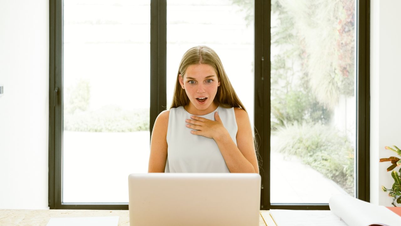 A female worker at her desk looks happily surprised while looking at her laptop.