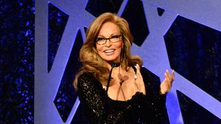 Raquel Welch clapping behind a podium. She's wearing glasses and a black sequenced dress.