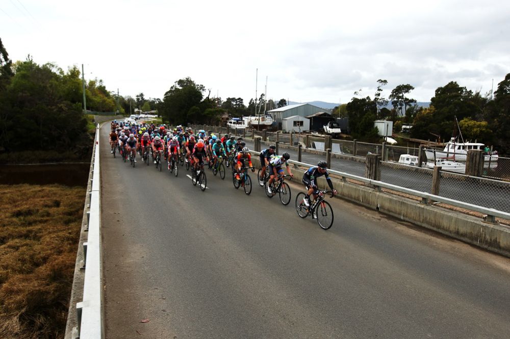 The peloton rolls past a small village on the Tamar river.