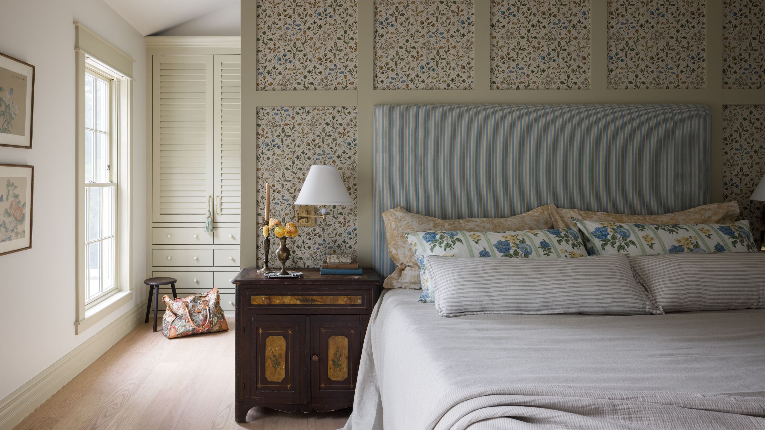 a large white and soft yellow beige painted bedroom with a central dividing wall with patterned framed insets, striped headboard, antique nightstand and closets behind