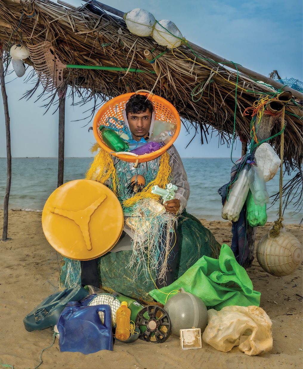 A man sits on a sandy beach under a thatched shelter, wearing a costume made from fishing nets, plastic bottles and an orange basket around his neck, surrounded