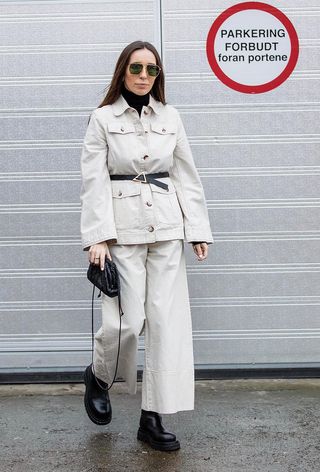 how to wear Chelsea boots is shown in a photo of a woman standing outside wearing a black turtleneck under a white jacket with matching white pants and black chelsea boots