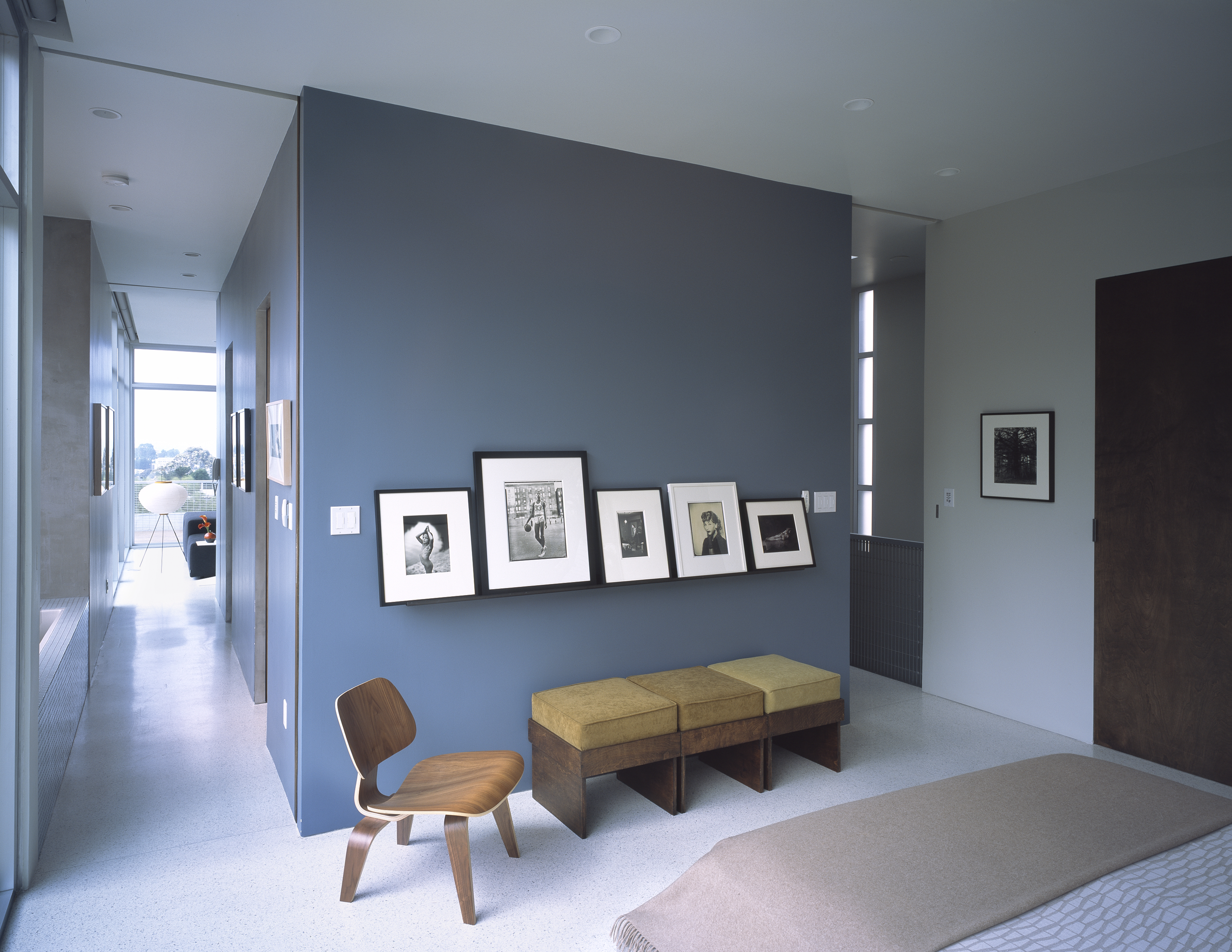 Blue bedroom with picture ledge holding photographs, bed, wooden bench with yellow cushions and midcentury wooden chair