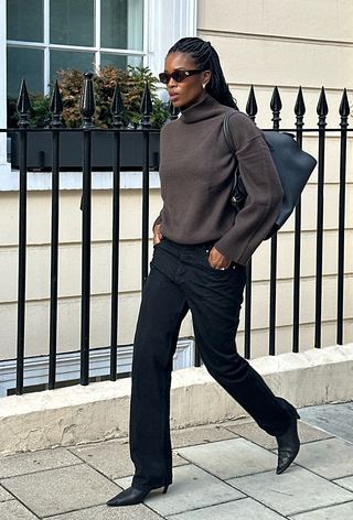 The winter capsule wardrobe item, turtleneck sweater, is shown in a photo of a woman walking down the sidewalk wearing brown sunglasses, a brown turtleneck sweater, black skinny jeans, black ankle boots, and a black handbag