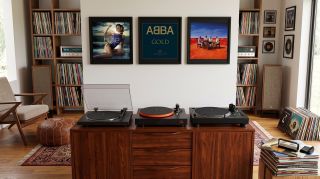 Three turntables on a media unit, with some records hanging on a wall behind.