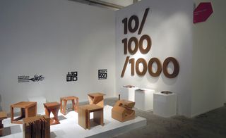 Wooden side tables of different shapes on a raised white platform in front of a wall with logos printed on it and a wall with the wooden numbers 10, 100 and 1000 on it.