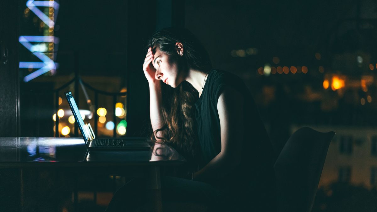 A stressed female IT worker, shot from the side in a dark room and lit by her laptop screen, holding her head in her hand to represent IT skill shortages.