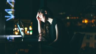 A stressed female IT worker, shot from the side in a dark room and lit by her laptop screen, holding her head in her hand to represent IT skill shortages.