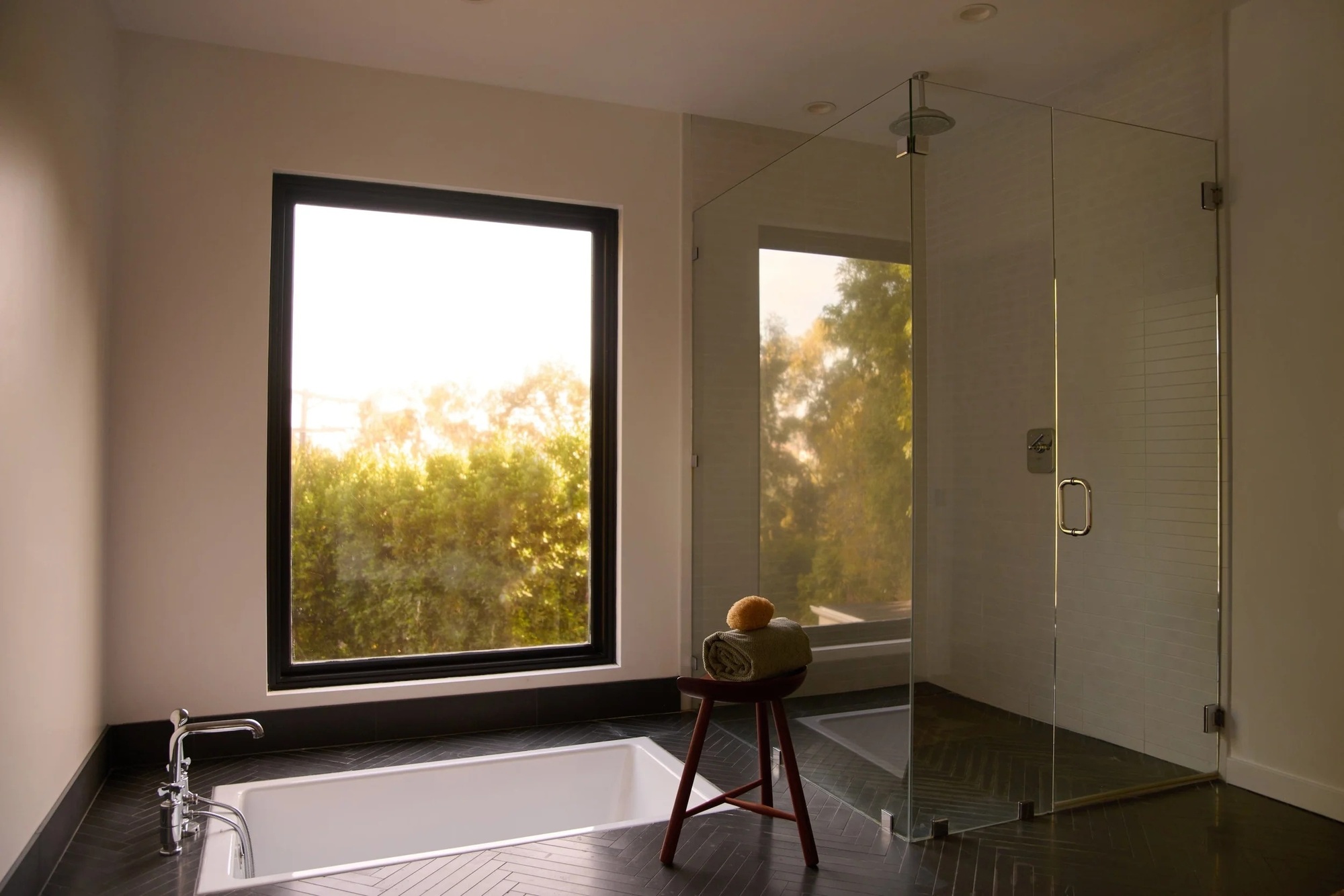 white bathroom with black tiled flooring. white sunken bath tub in front of large picture window. on the right is glass walk-in shower