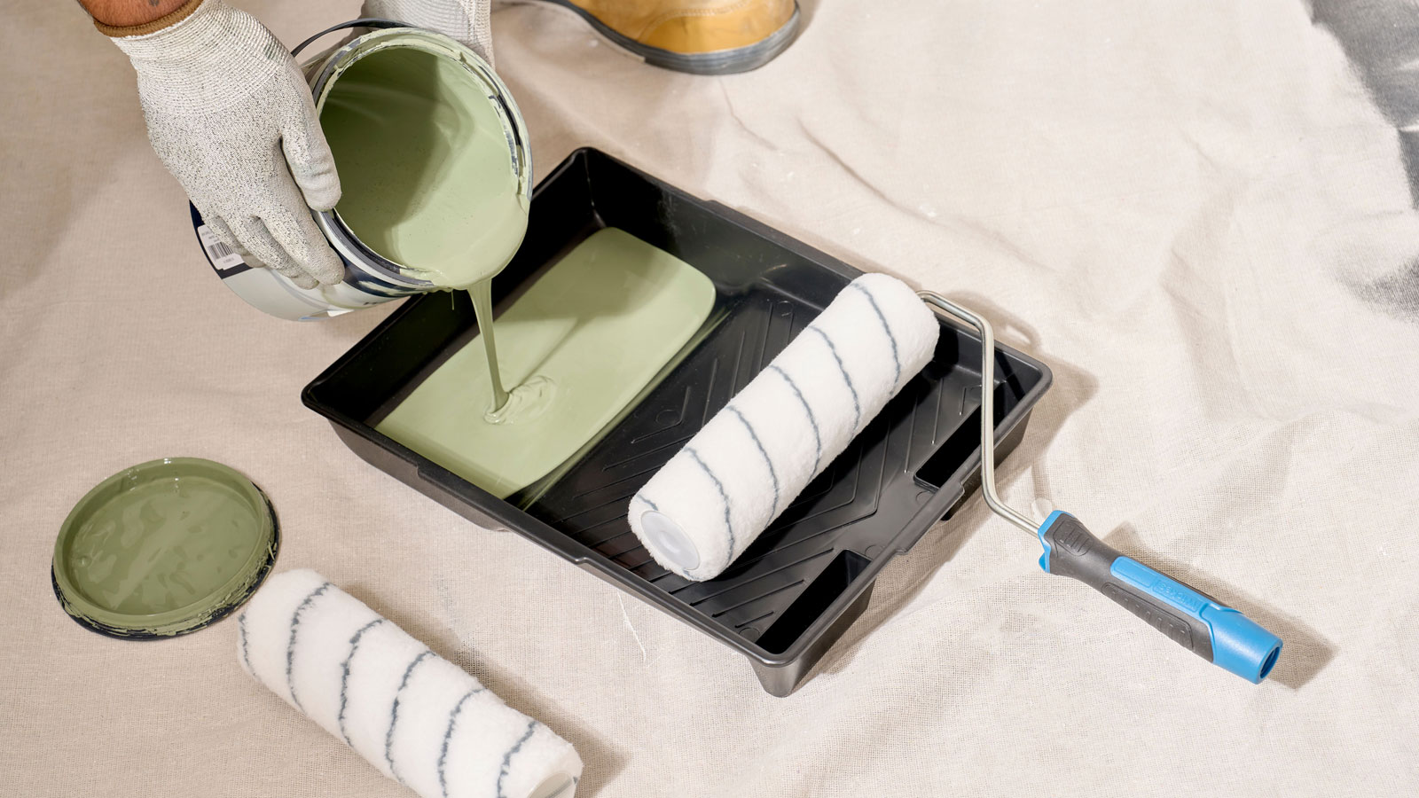 Person pouring green paint from tin into roller tray with roller resting on tray, all placed on a dust sheet