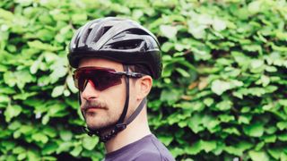 A man in a bike helmet and sunglasses in front of a hedge
