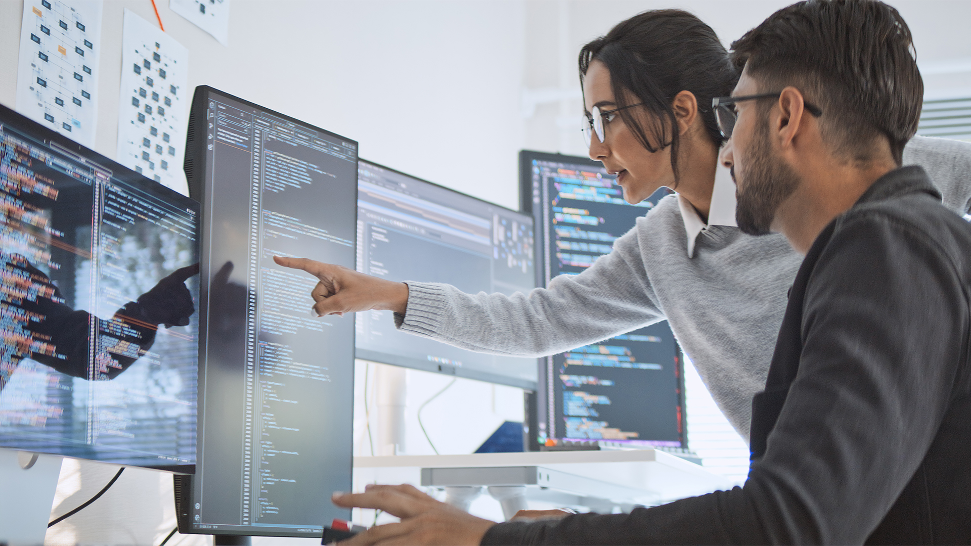 Female forward deployed engineer standing over the shoulder of male software engineer offering them practical advice on code while pointing at multi-monitor setup on desk.