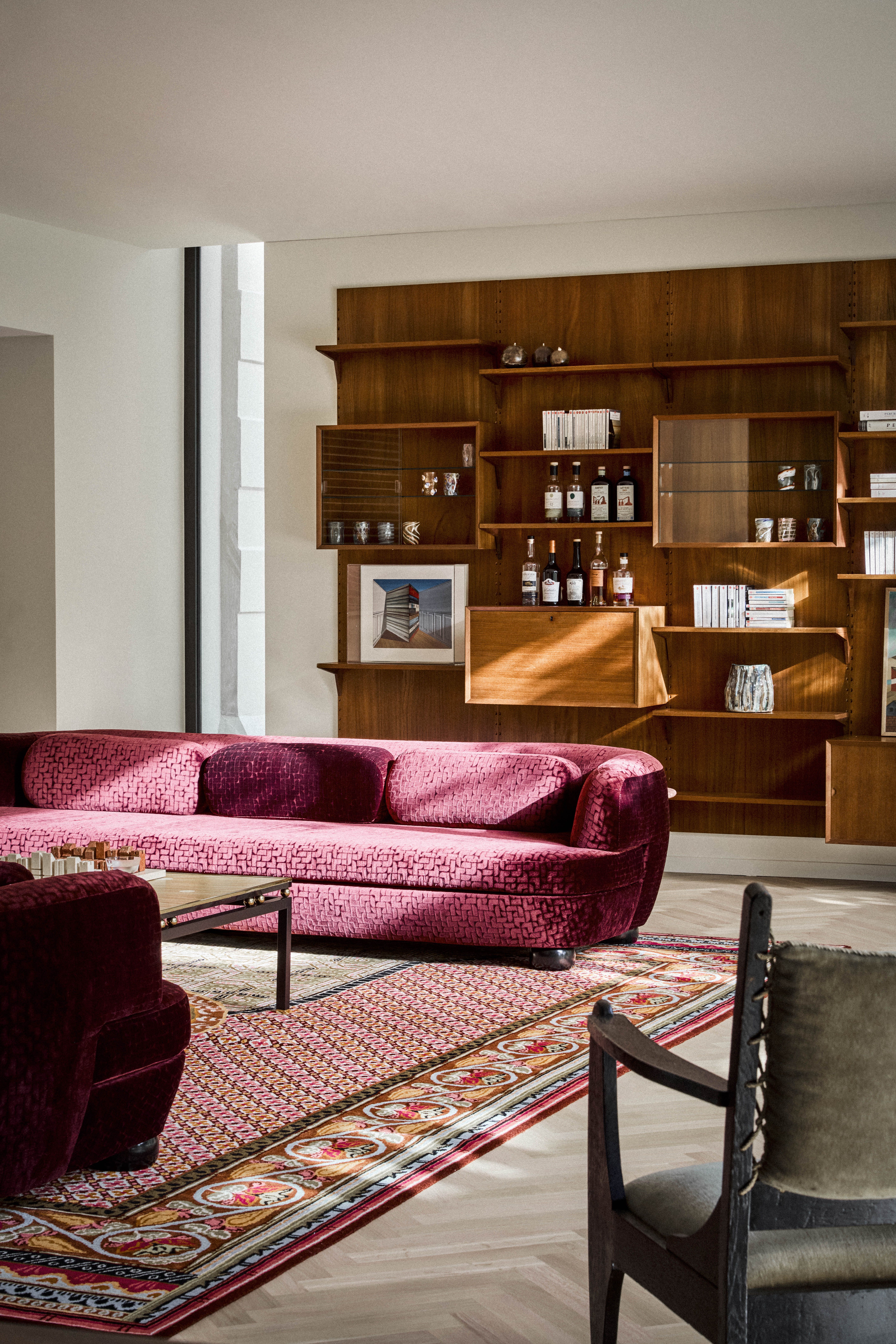 A living room with a red printed rug, a deep toned velvet sofa, an armchair, a standing lamp and a bookshelf 