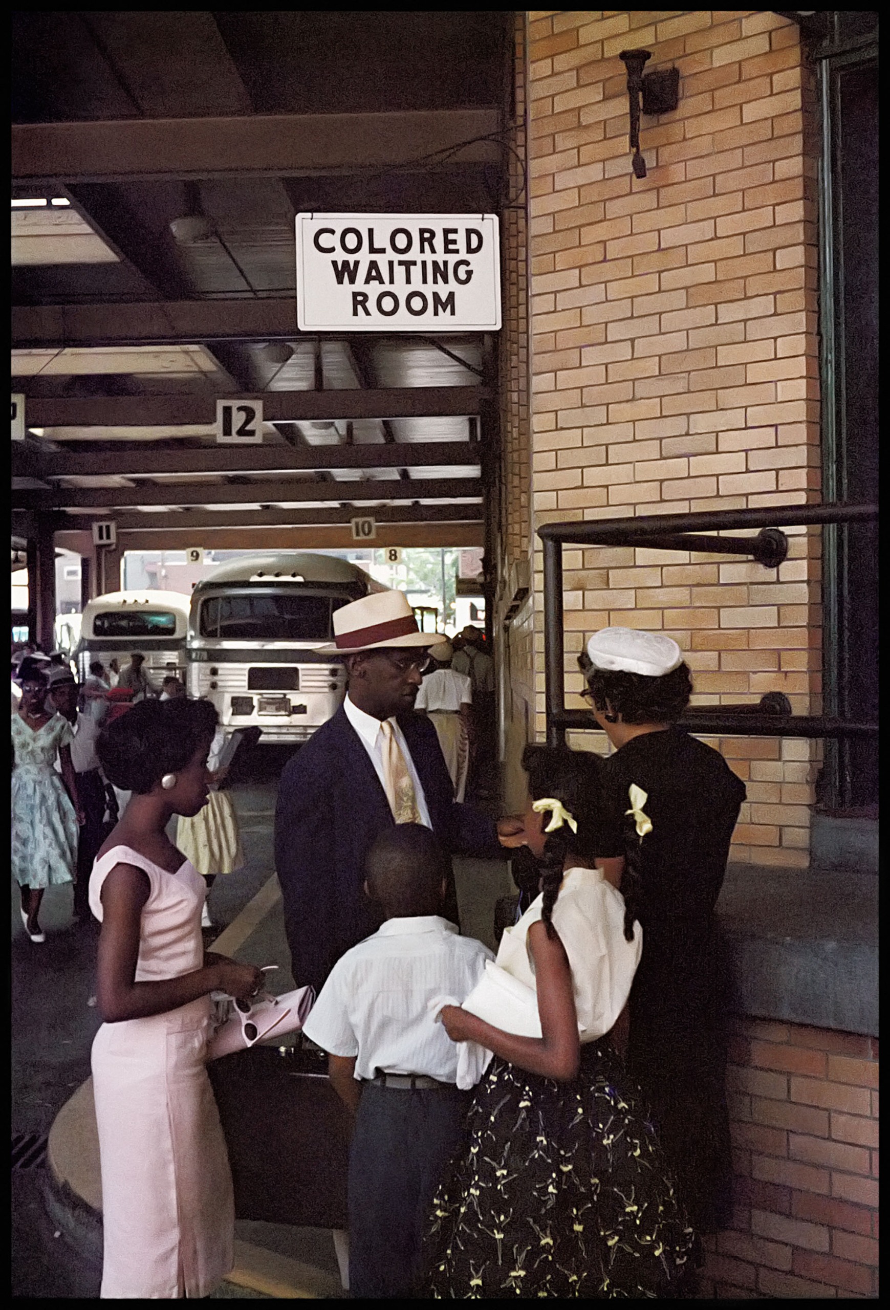 A sign reading "Colored Waiting Room" hangs above a group of Black travellers at a segregation-era bus station, with buses idling in the background
